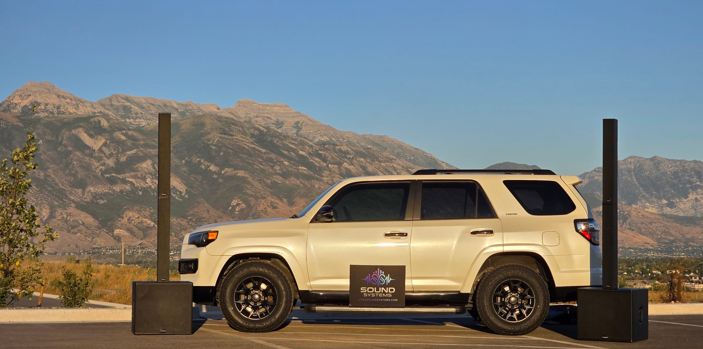 A white Sound Systems SUV parked below the Wasatch mountains, with column speakers on subwoofers set up beside it