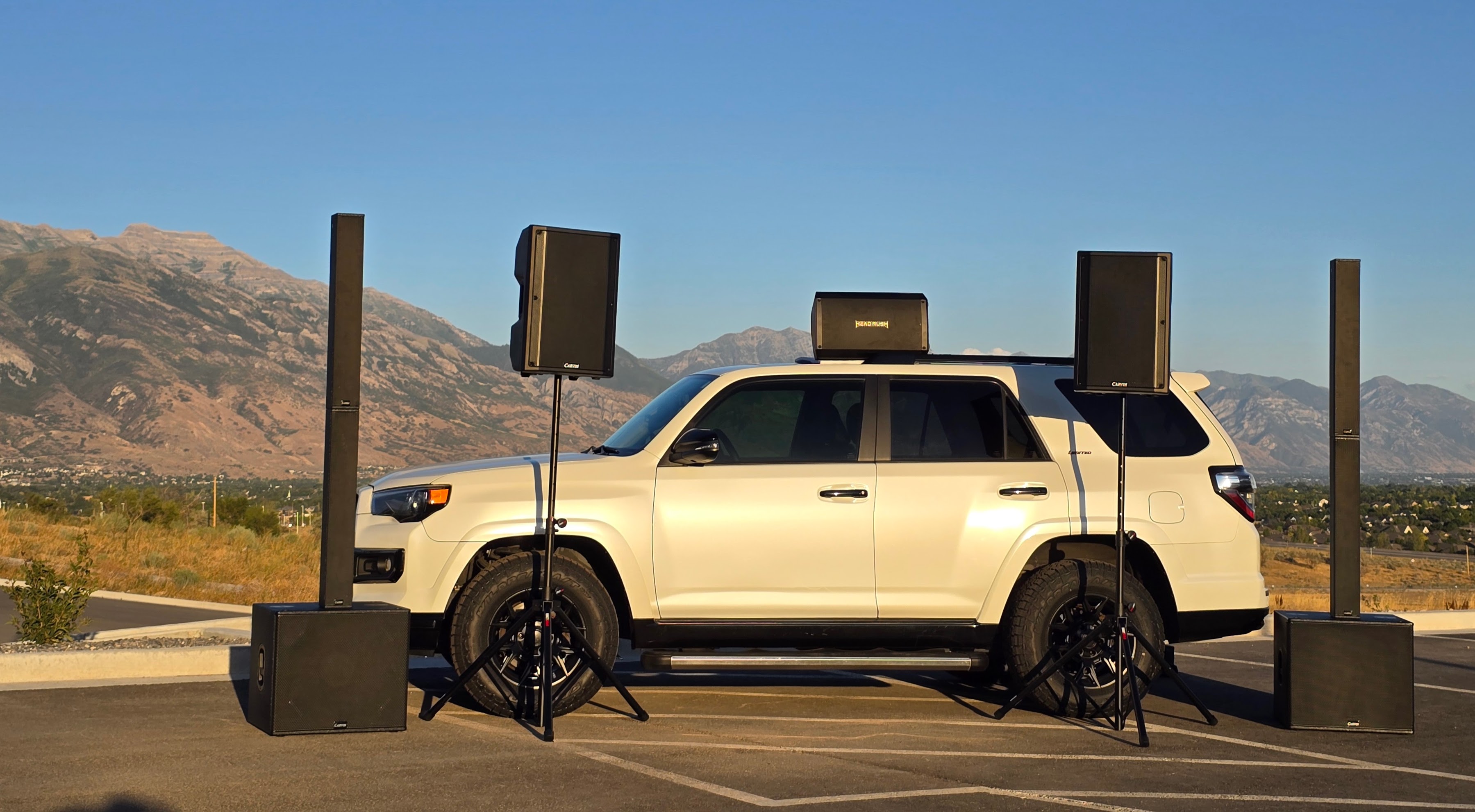 A full event system staged around a white Toyota 4Runner at golden hour: two column-array towers, two Carvin speakers on tripod stands, two subwoofers on the ground, and a rack unit on the roof, with the Wasatch mountains behind